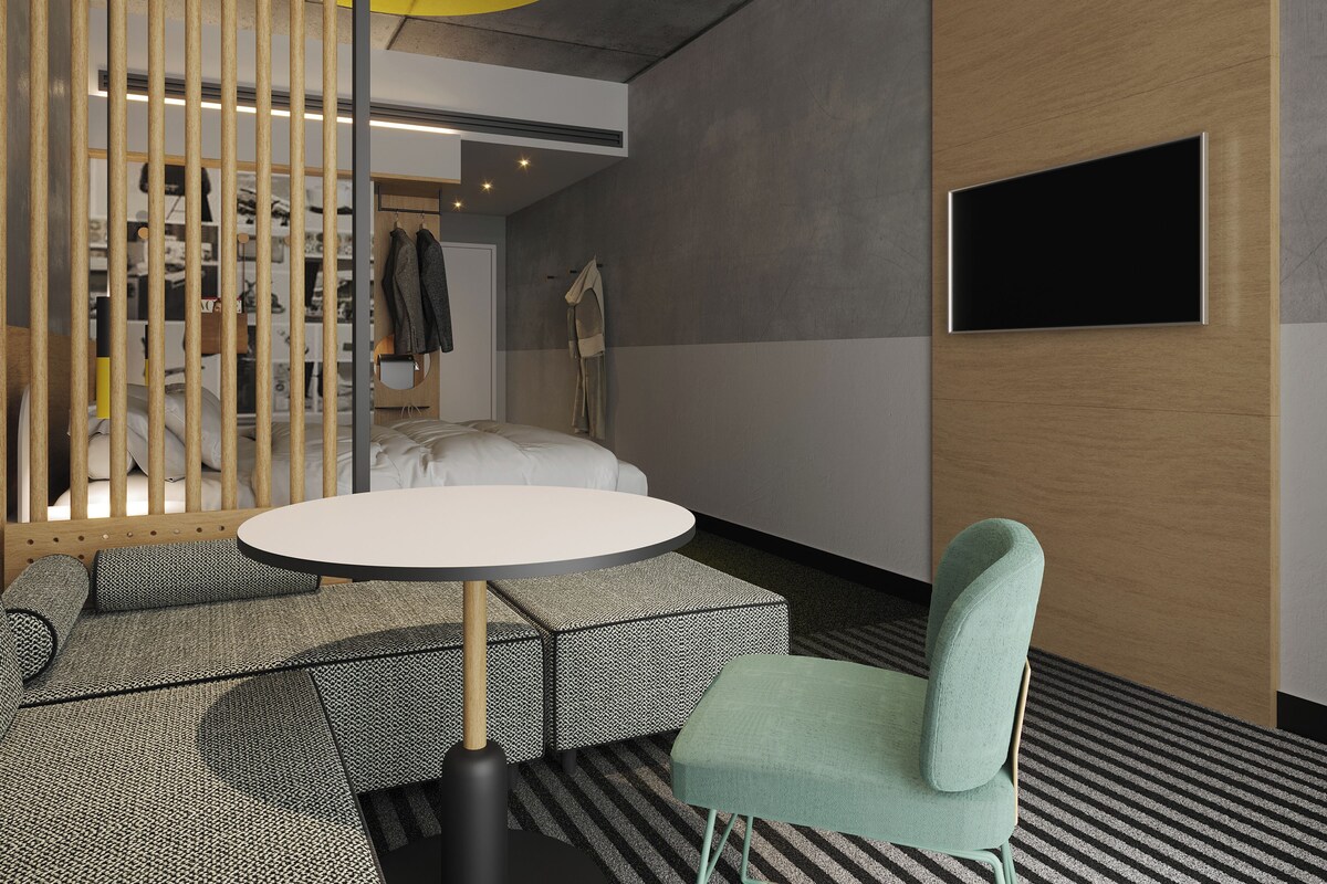 Una habitación moderna con una mesa redonda, una silla verde, una cama con cabecera, un armario y un televisor montado en la pared.