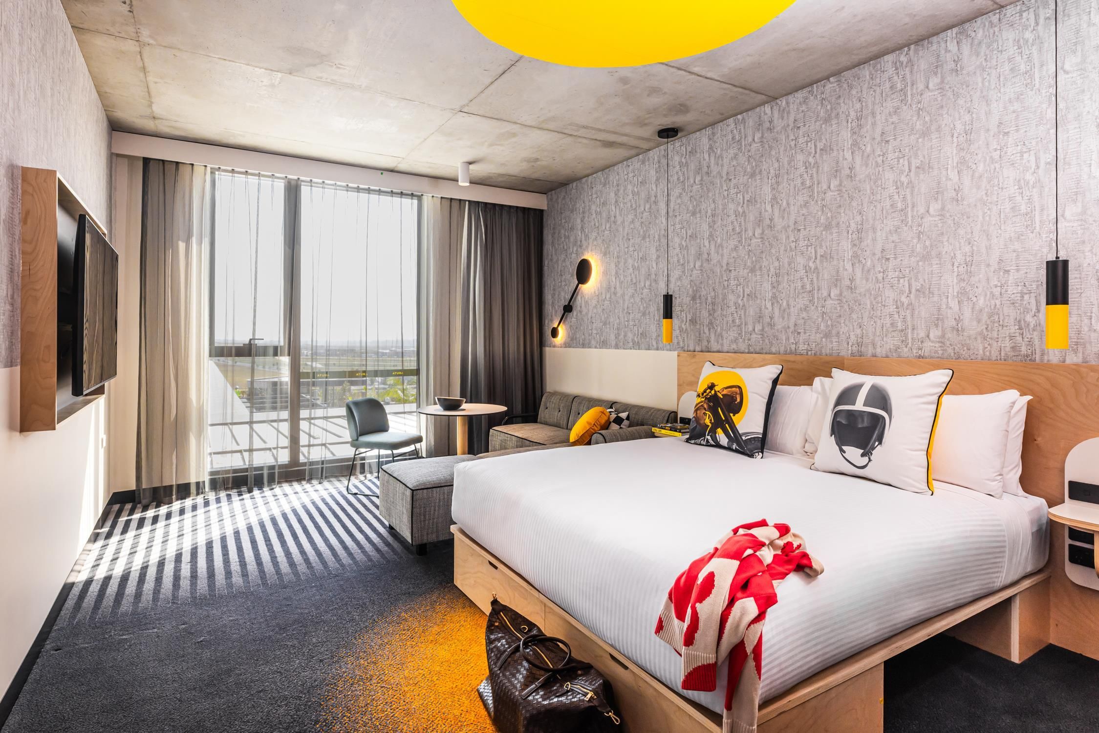 Habitación de hotel con una cama grande, un escritorio, una silla, una mesita y vistas de la ciudad a través de amplios ventanales.