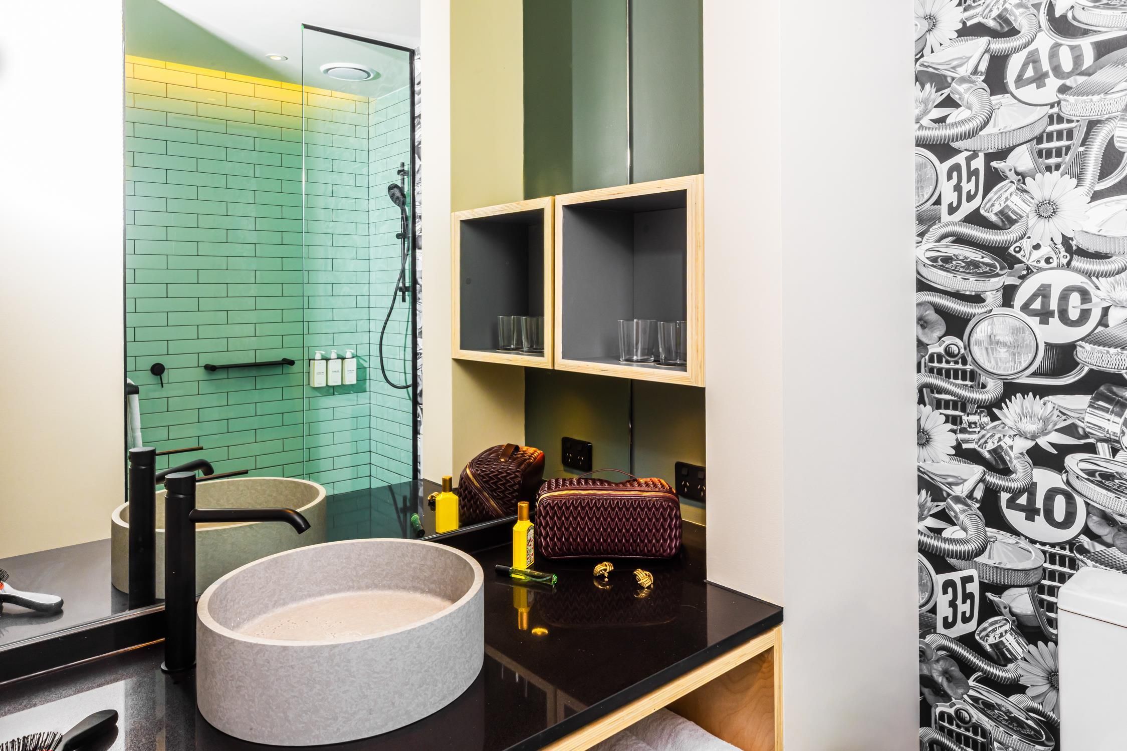 A bathroom with a green-tiled wall, a black countertop, a round basin, and two shelves with glasses and a handbag.