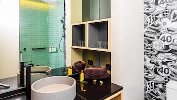 A bathroom with a green-tiled wall, a black countertop, a round basin, and two shelves with glasses and a handbag.