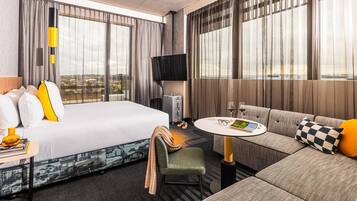 A hotel room with a bed, a sofa, a small table, a TV, and a view of the city.