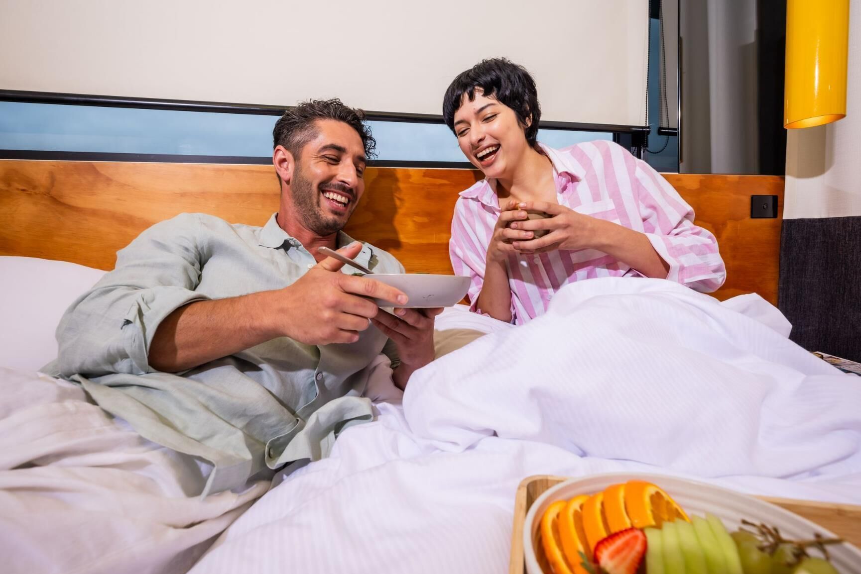 Dos personas en una cama con una tablet, una mesita de noche con frutero y un cabecero de madera.