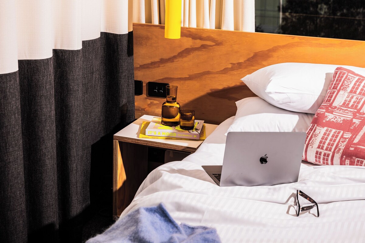 Una cama bien hecha con una computadora portátil, un par de gafas y un libro titulado 'It's a Mood' sobre una mesita de noche.