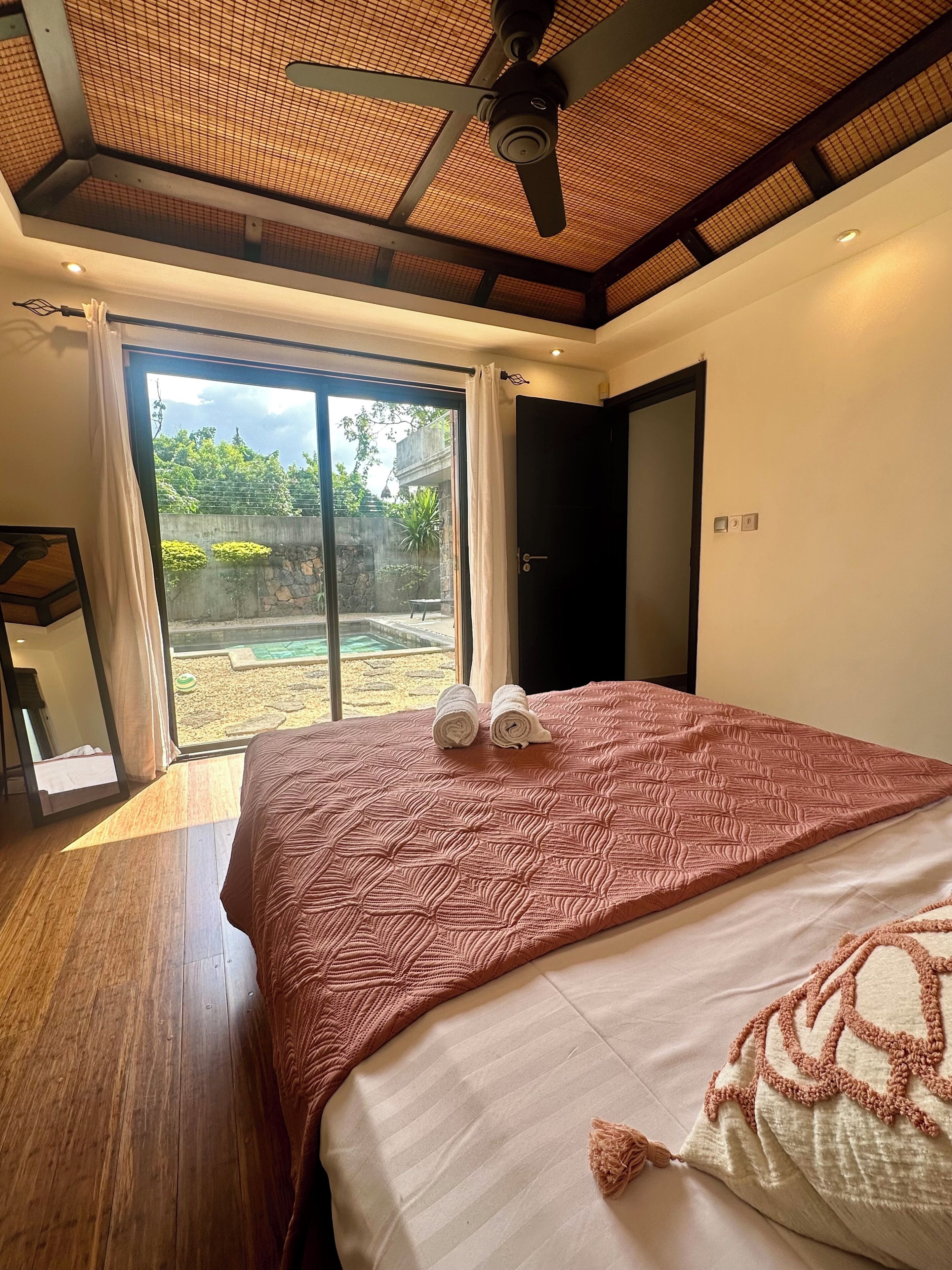 A bedroom with a large bed, a ceiling fan, a sliding glass door to a courtyard, and a dark door.