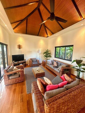 Ein geräumiges Wohnzimmer mit Rattanmöbeln, Deckenventilator und großen Fenstern.
