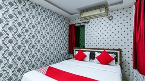 Una cama bien tendida con un detalle rojo, un papel tapiz estampado, una ventana con cortinas rojas y un equipo de aire acondicionado.