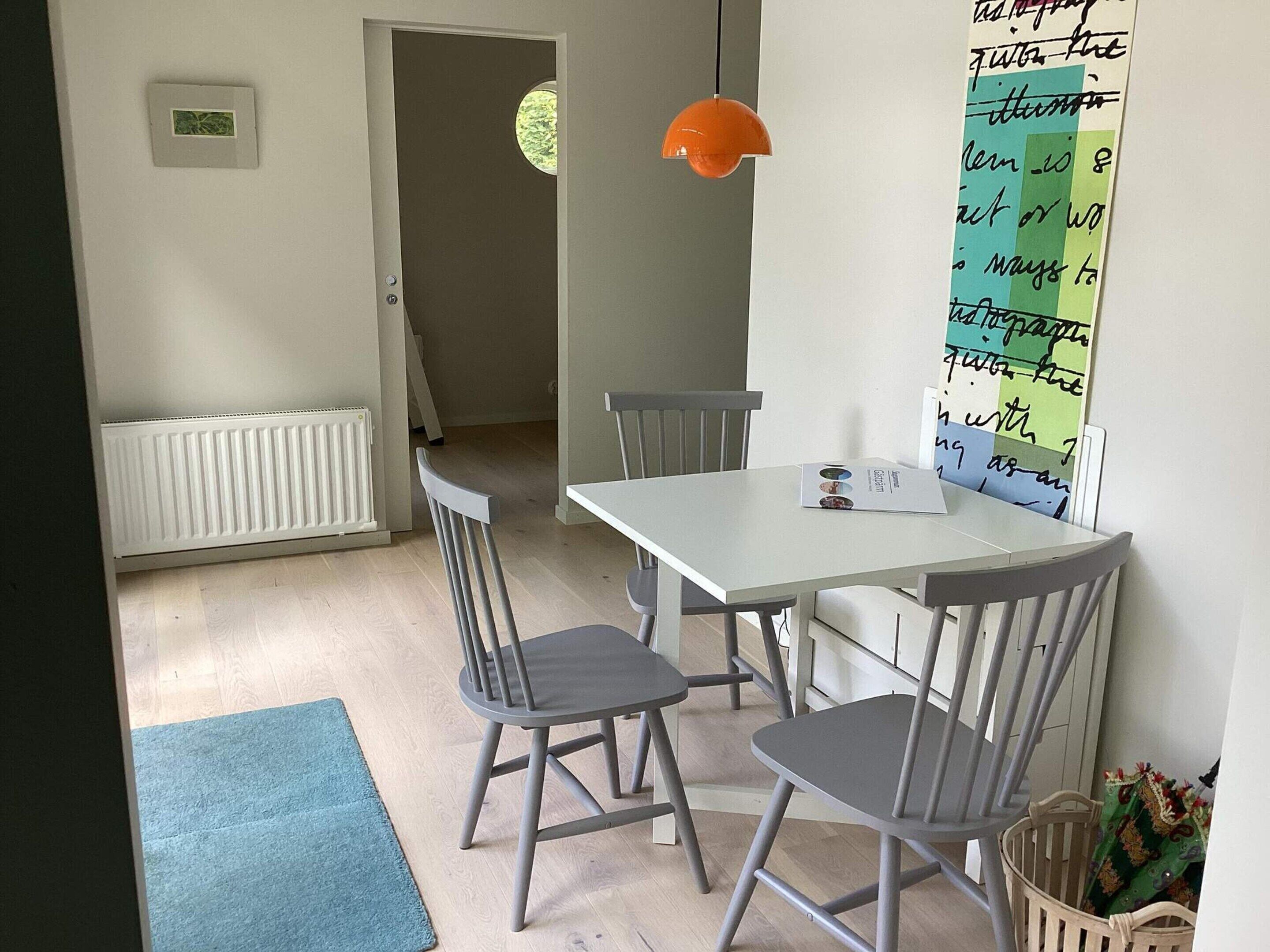 A dining area with a white table, four grey chairs, a blue rug, a hanging orange pendant light, and a wall with a green and white poster.