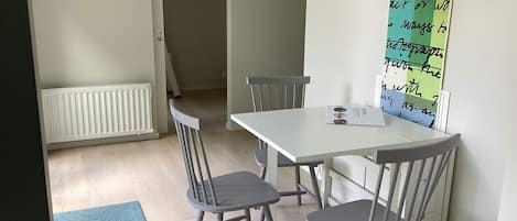 A dining area with a white table, four grey chairs, a blue rug, a hanging orange pendant light, and a wall with a green and white poster.