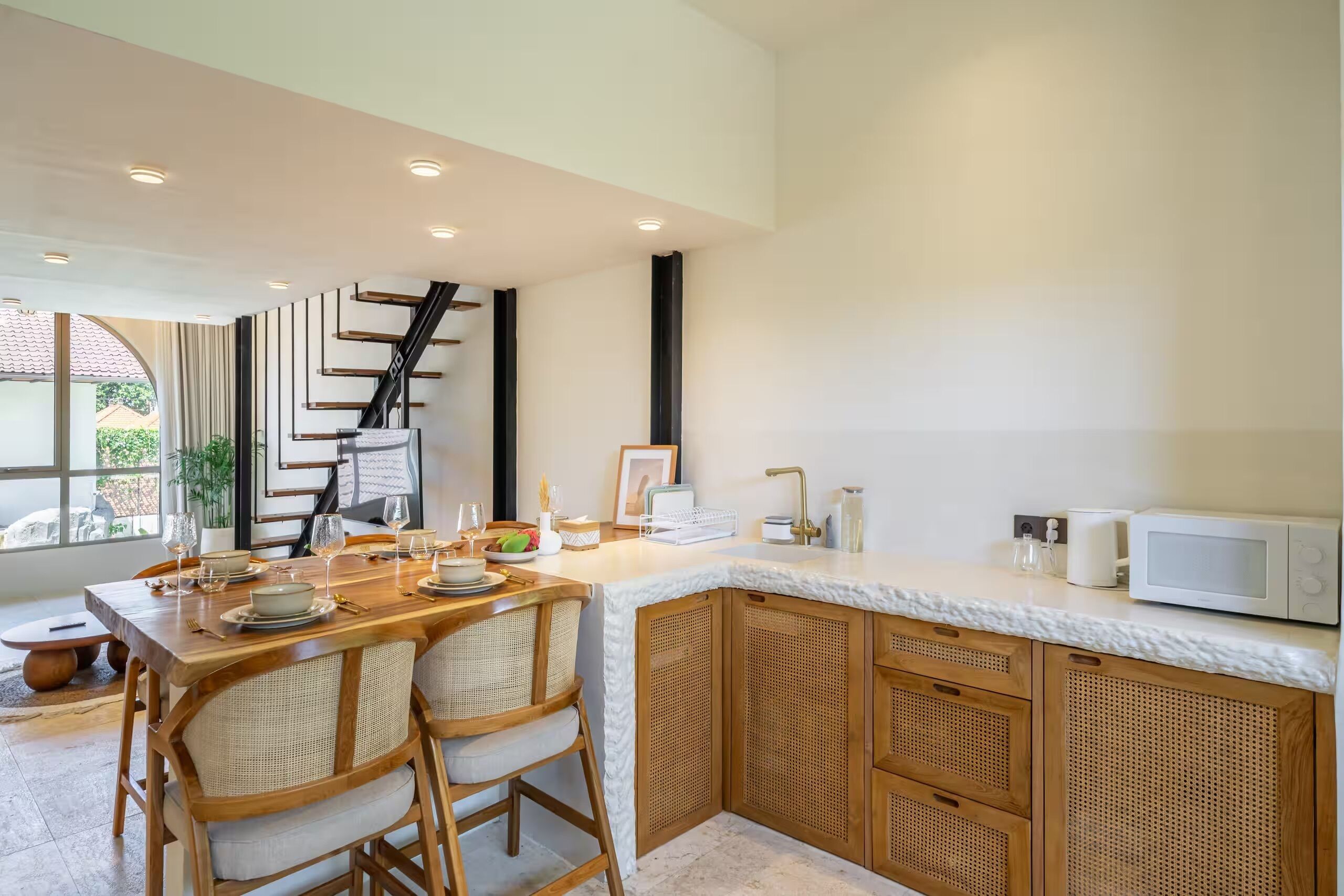 A modern kitchen with a dining area, featuring a wooden table set for four, a white countertop, and a staircase leading to another level.