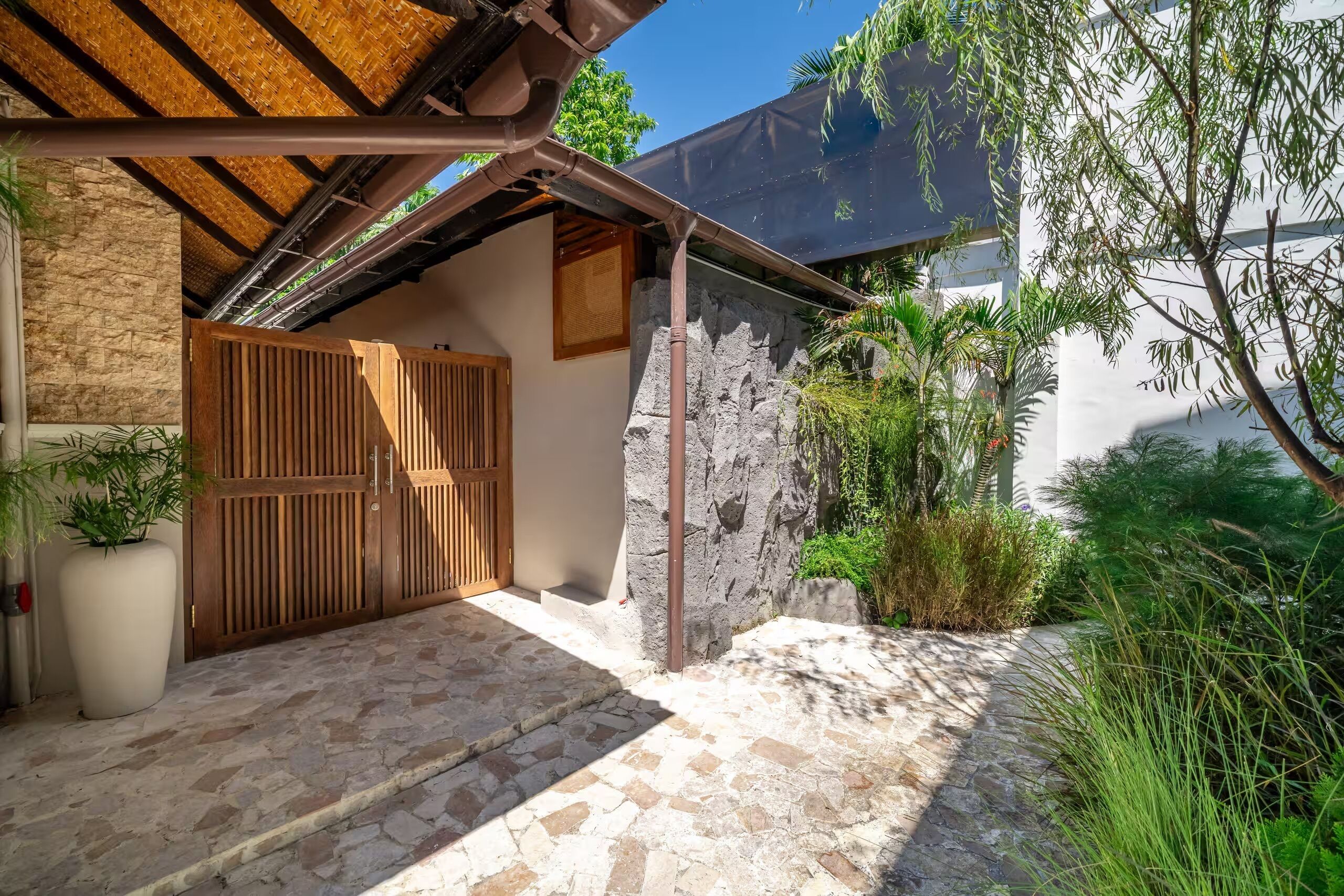 A stone-paved entrance with a wooden gate, a thatched roof, and a white wall with a stone facade.
