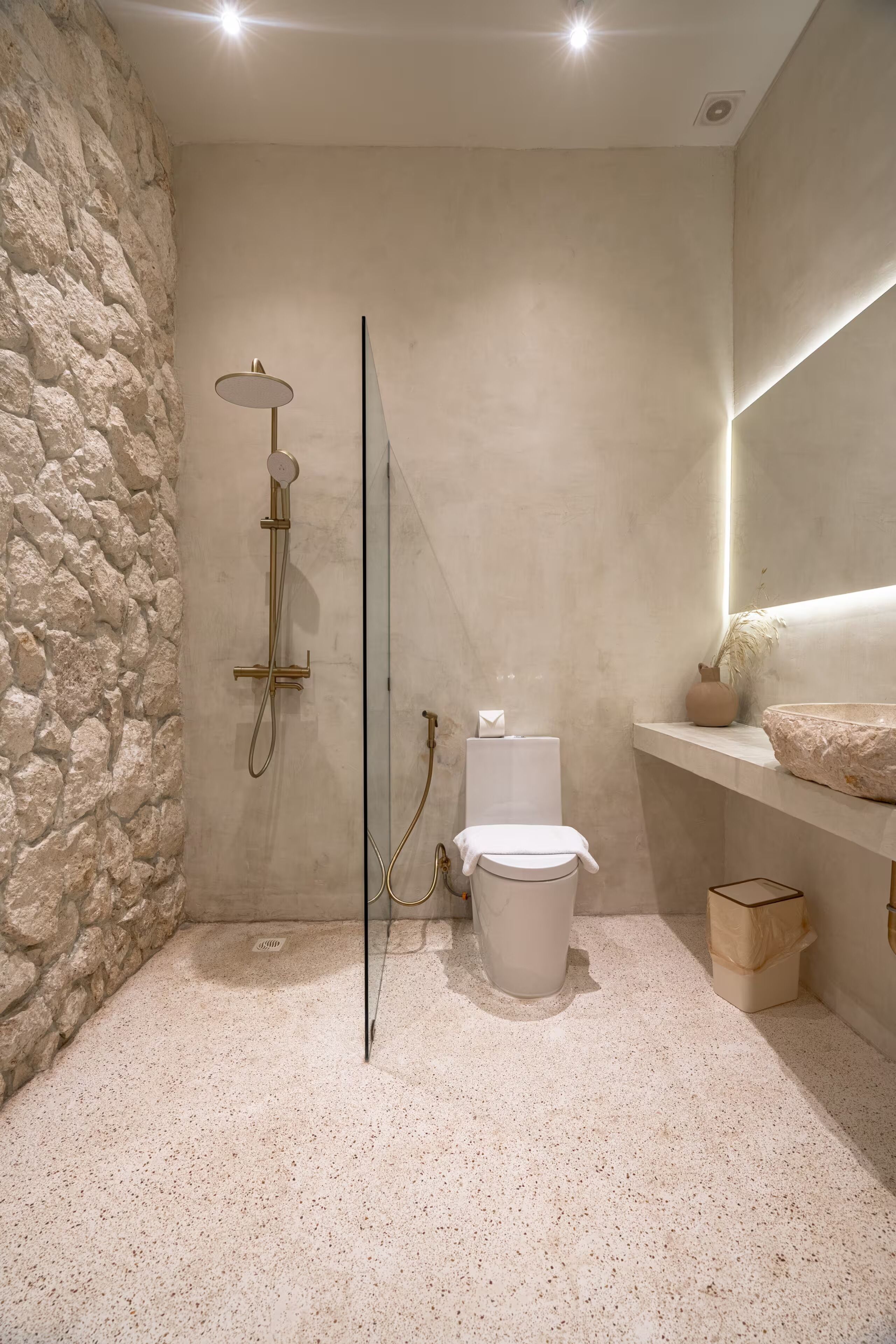 A bathroom with a stone wall, a white toilet, a shower with a golden showerhead, and a shelf with a vase and a bowl.