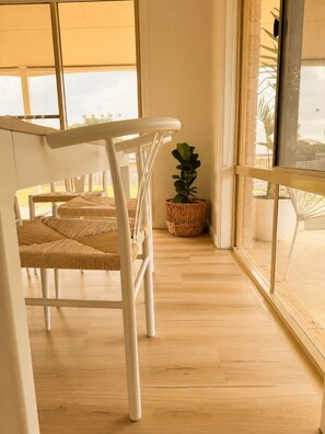 A room with a white chair, a woven table, a wicker basket, and a view through large windows.