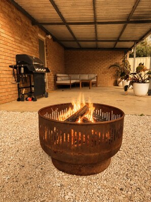 A fire pit in a covered outdoor area with a brick wall, a grill, a sofa, and potted plants.