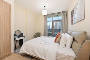 A modern hotel room with a bed, a desk, a chair, a city view, and a hanging light fixture. - Bloomfields 2BR  Canal Bay Business Bay (Dubai)
