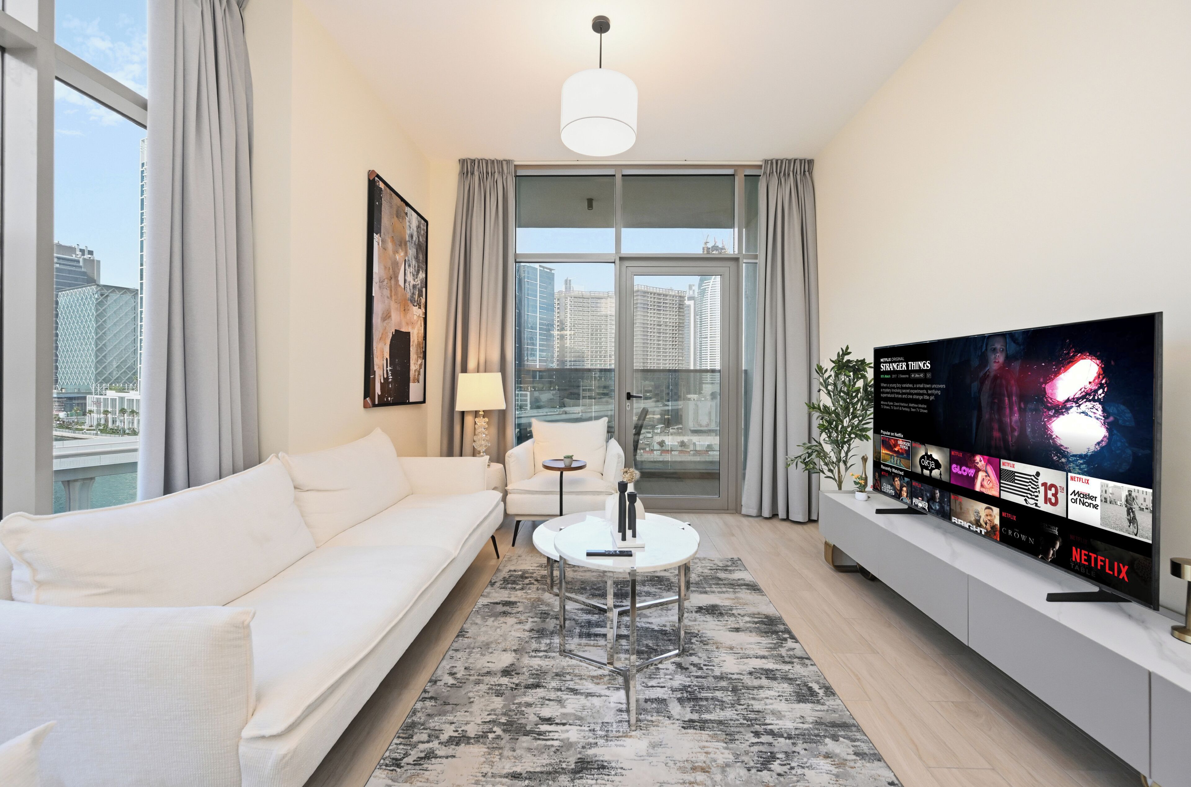 Una sala de estar moderna con un sofá blanco, una mesa de centro redonda, un televisor sobre un mueble y vistas a la ciudad a través de amplias ventanas.