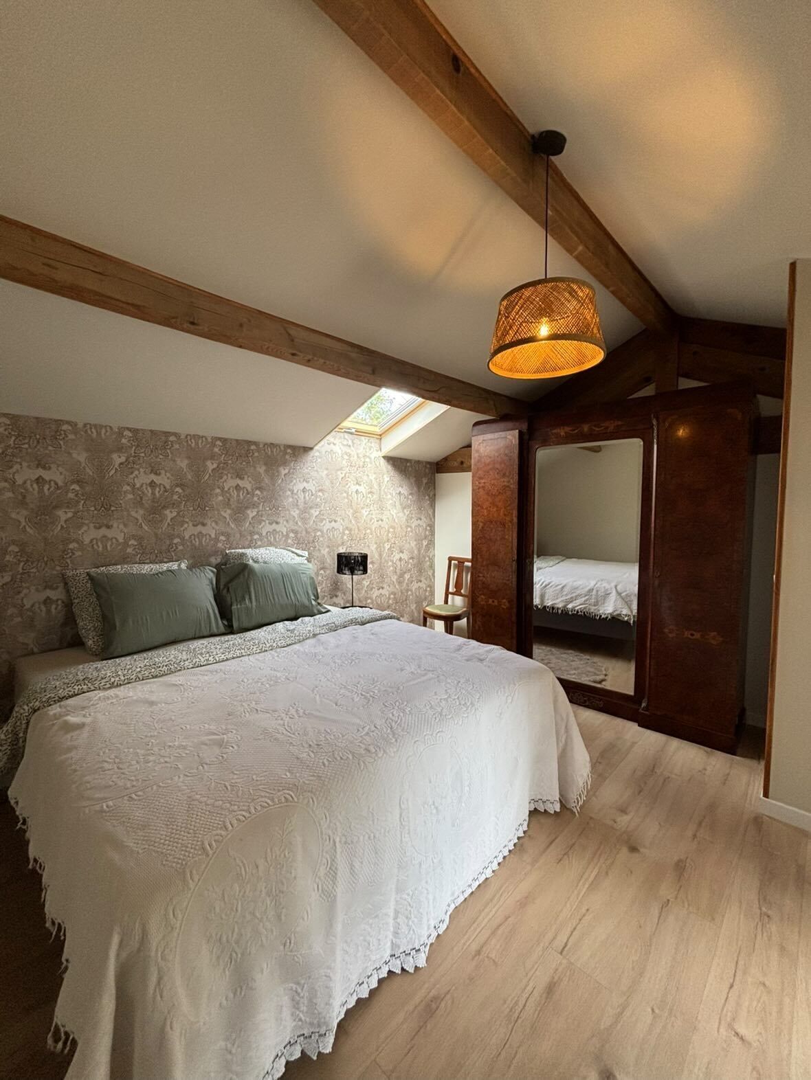 A bedroom with a large bed, wooden beams, a skylight, a hanging light fixture, and a mirror.