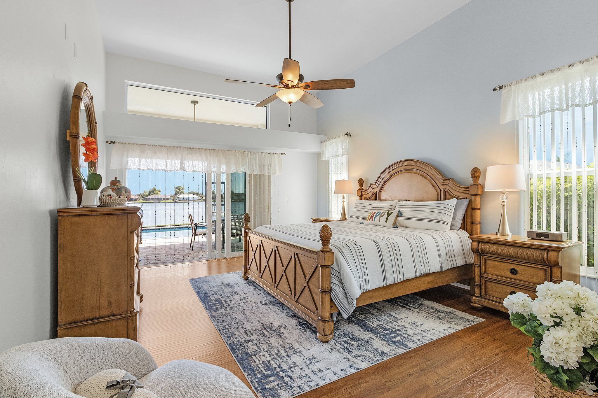 A bedroom with a bed, bedside tables, a ceiling fan, a dresser, a window with curtains, and a view of the outdoors.