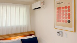 Quarto de hotel com cama, ar-condicionado, janela com cortinas leves, uma obra de arte emoldurada na parede e um termostato fixado na parede.