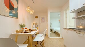 Cozinha moderna com armários brancos, sala de jantar com mesa posta para dois e quarto visível com uma cama.