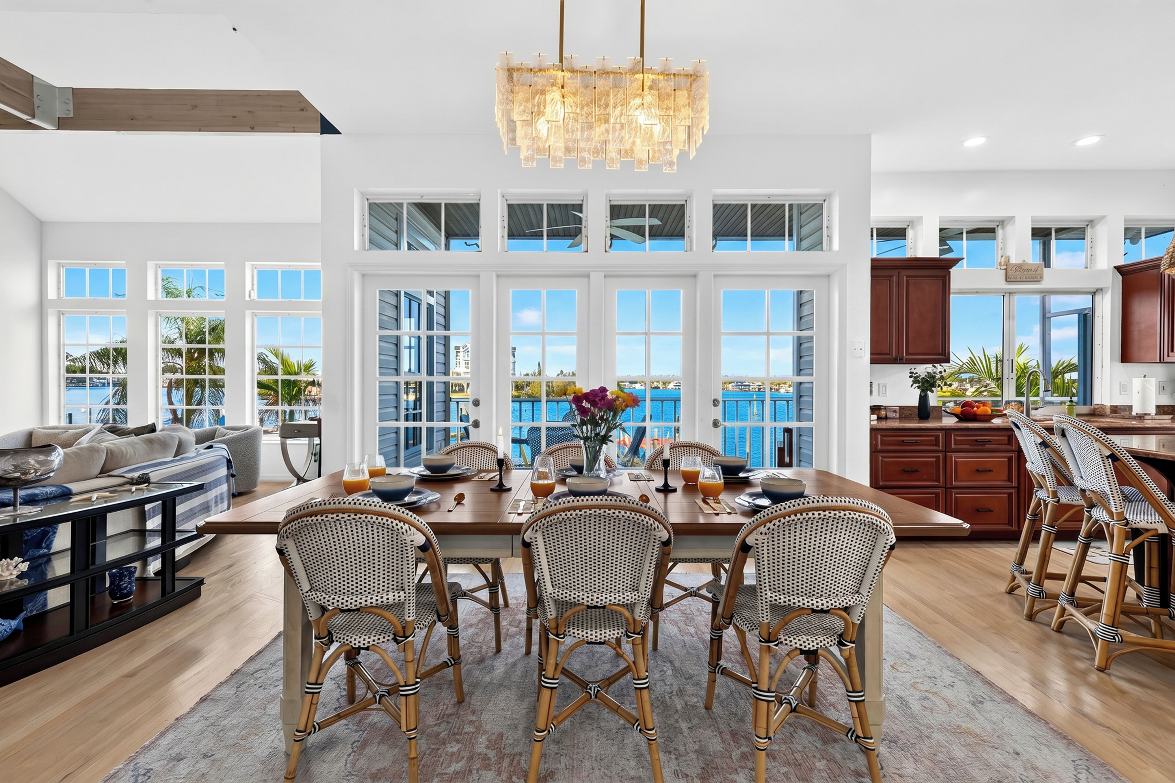A spacious kitchen with large windows, a dining area with wicker chairs, and a chandelier.