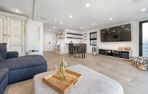 Ein modernes Wohnzimmer mit einer Couch, einem Couchtisch mit Weingläsern, einem Fernseher und einer Küche im Hintergrund.