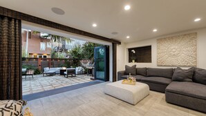 A modern living room with a grey sofa, a small ottoman, and a dining area with outdoor seating. - Rooftop Deck + Firepit + Wet Bar | Walk to Beach + Trail | Saltaire (Marina Del Rey)
