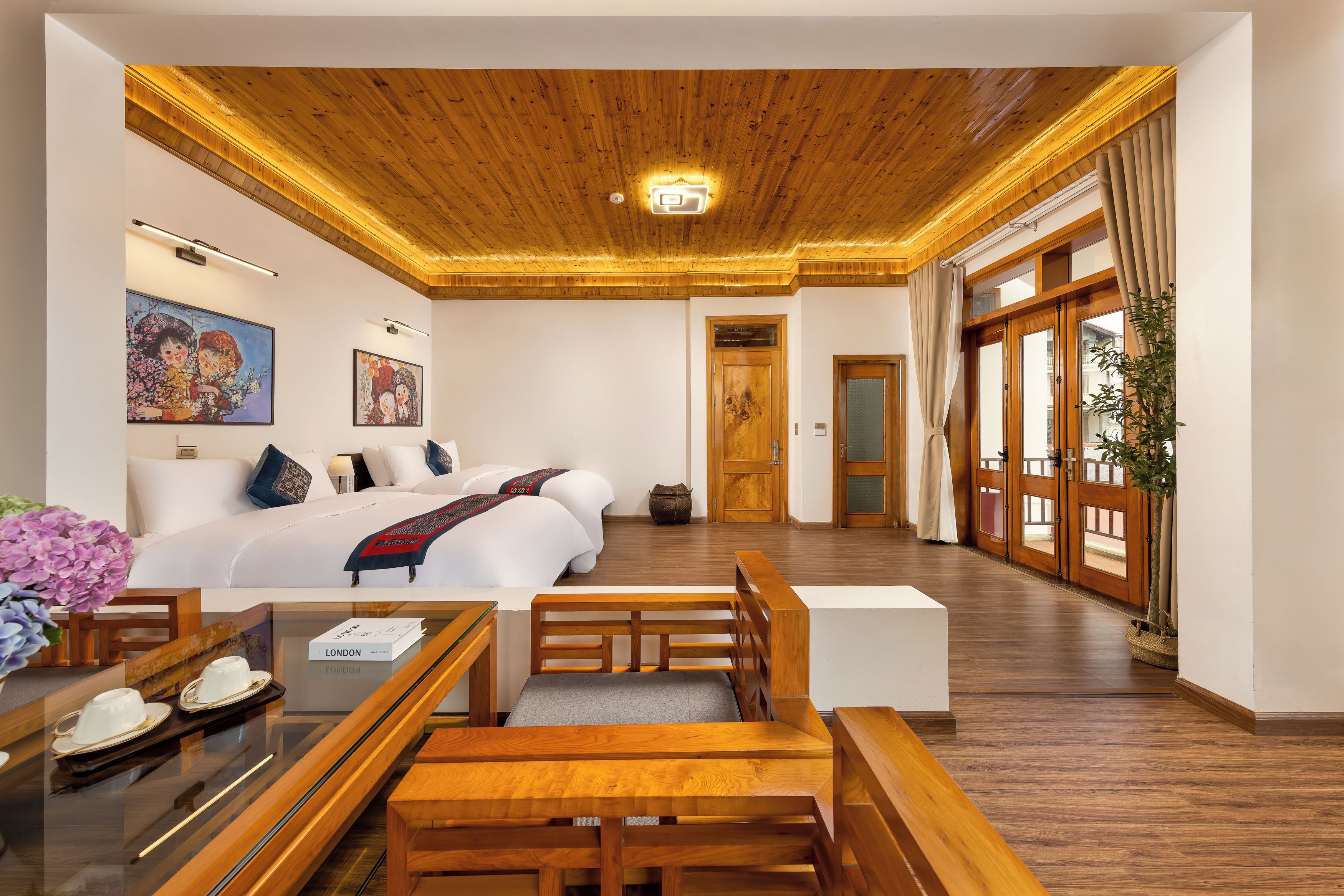Quarto de hotel com teto de madeira, cama com roupa de cama branca, escrivaninha com cadeira e uma janela grande com cortinas.
