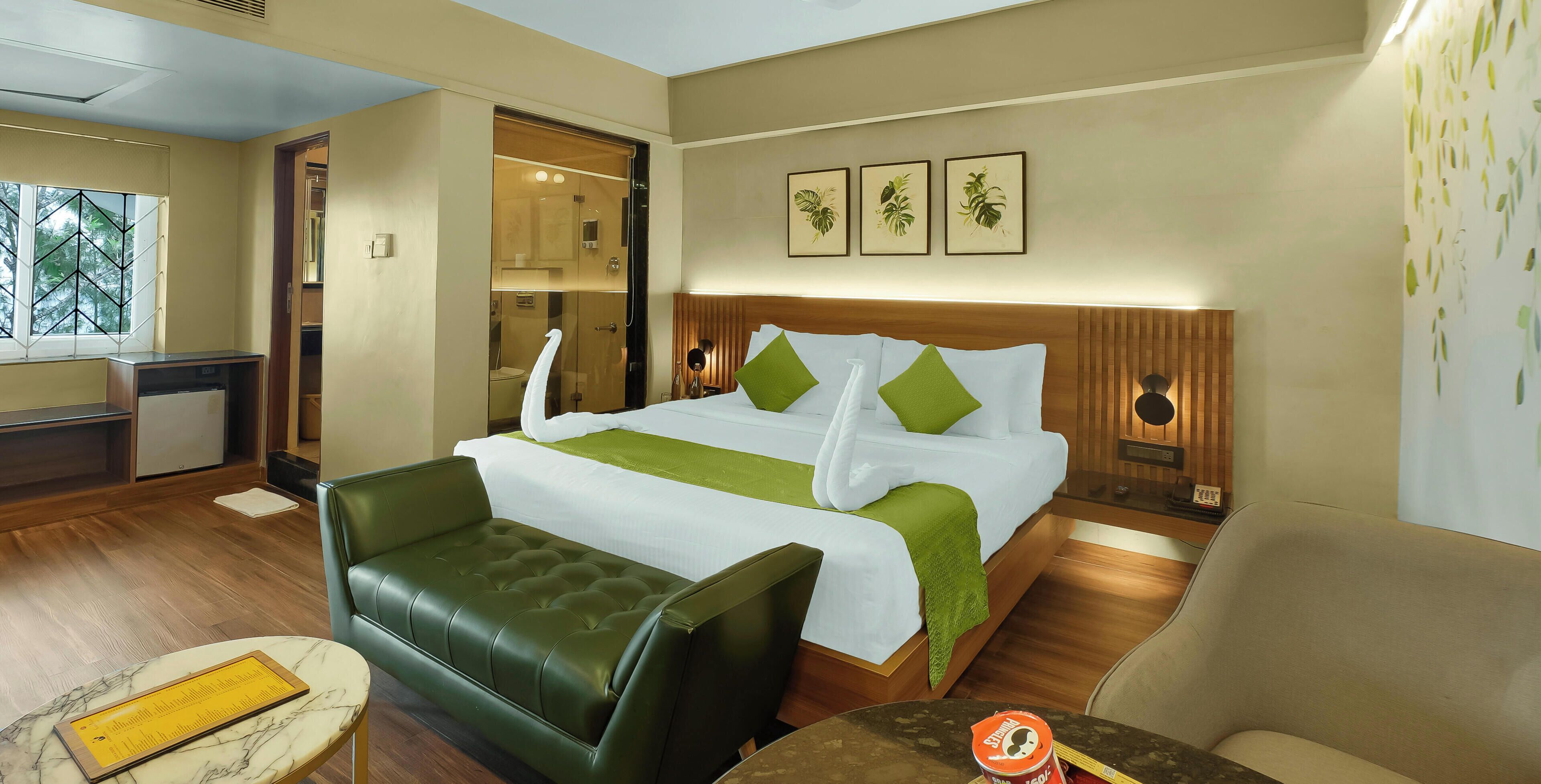 Ein Hotelzimmer mit Bett, einem grünen Sofa, einem kleinen Tisch, einem Spiegel und einem Fenster mit Blick auf Bäume.