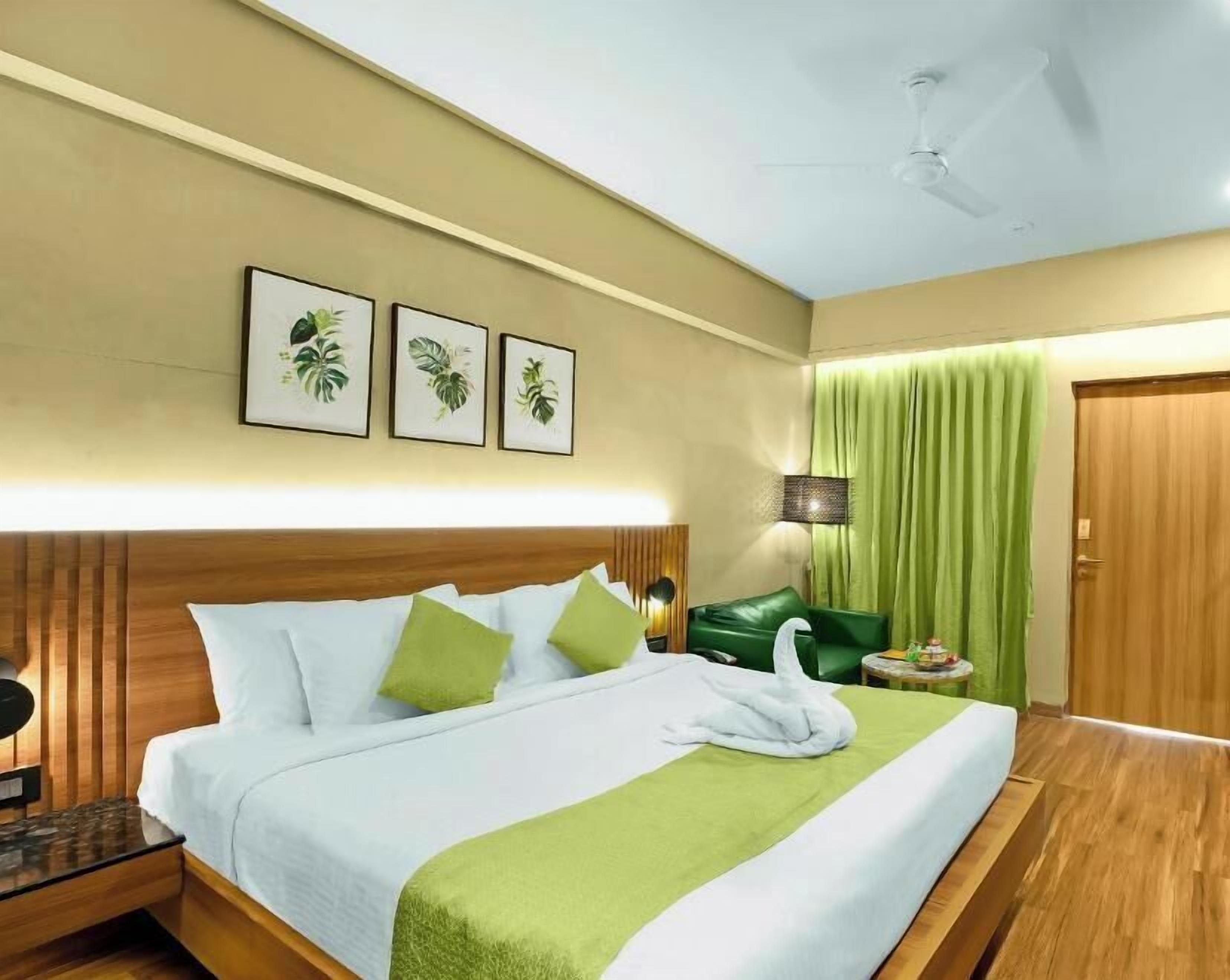 Een hotelkamer met een bed, nachtkastje, lamp, groen gordijn en lijstjes aan de muur.