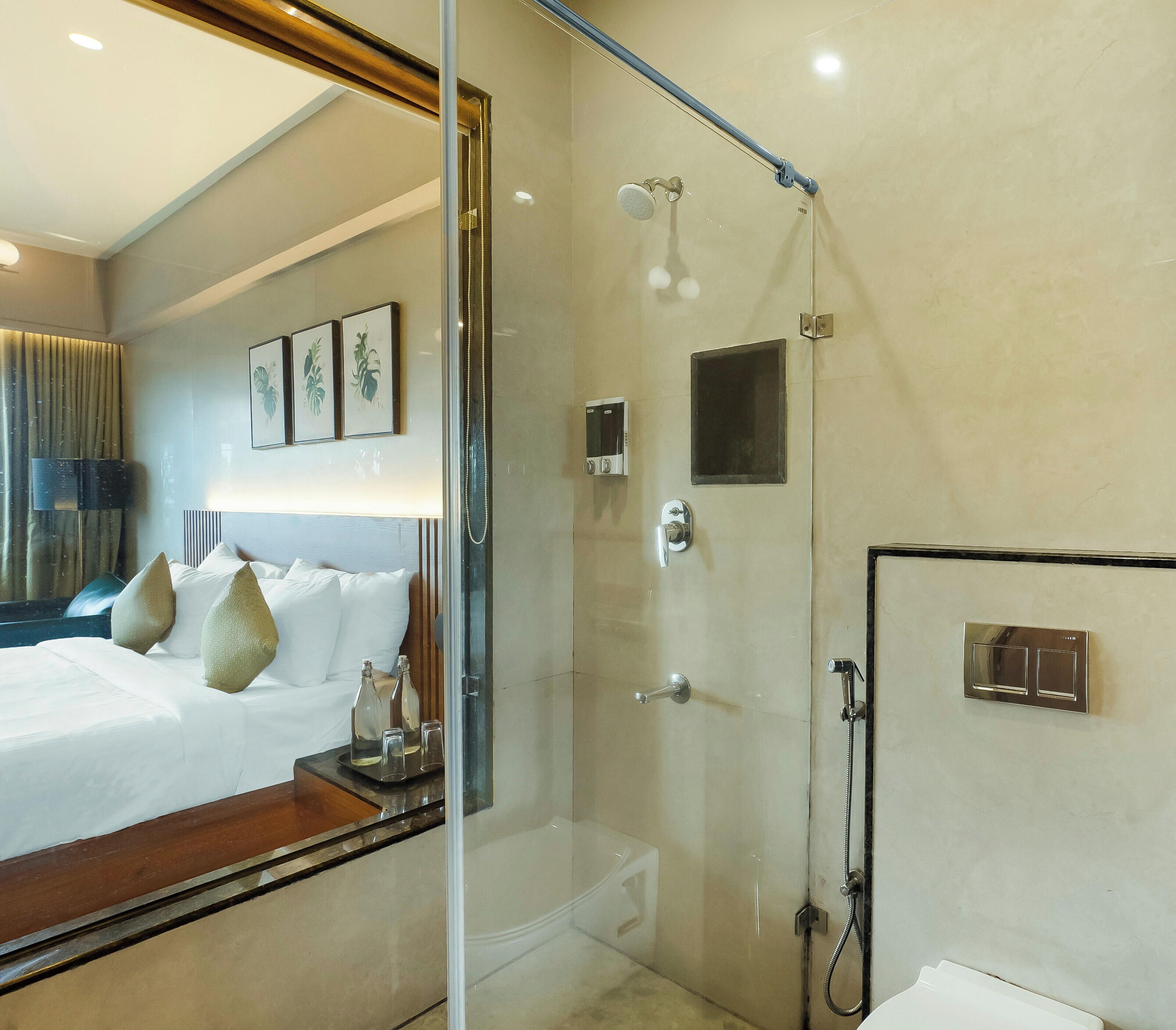 A hotel room with a glass-enclosed shower, a bed with white linens, and a bedside table with a bottle and glasses.