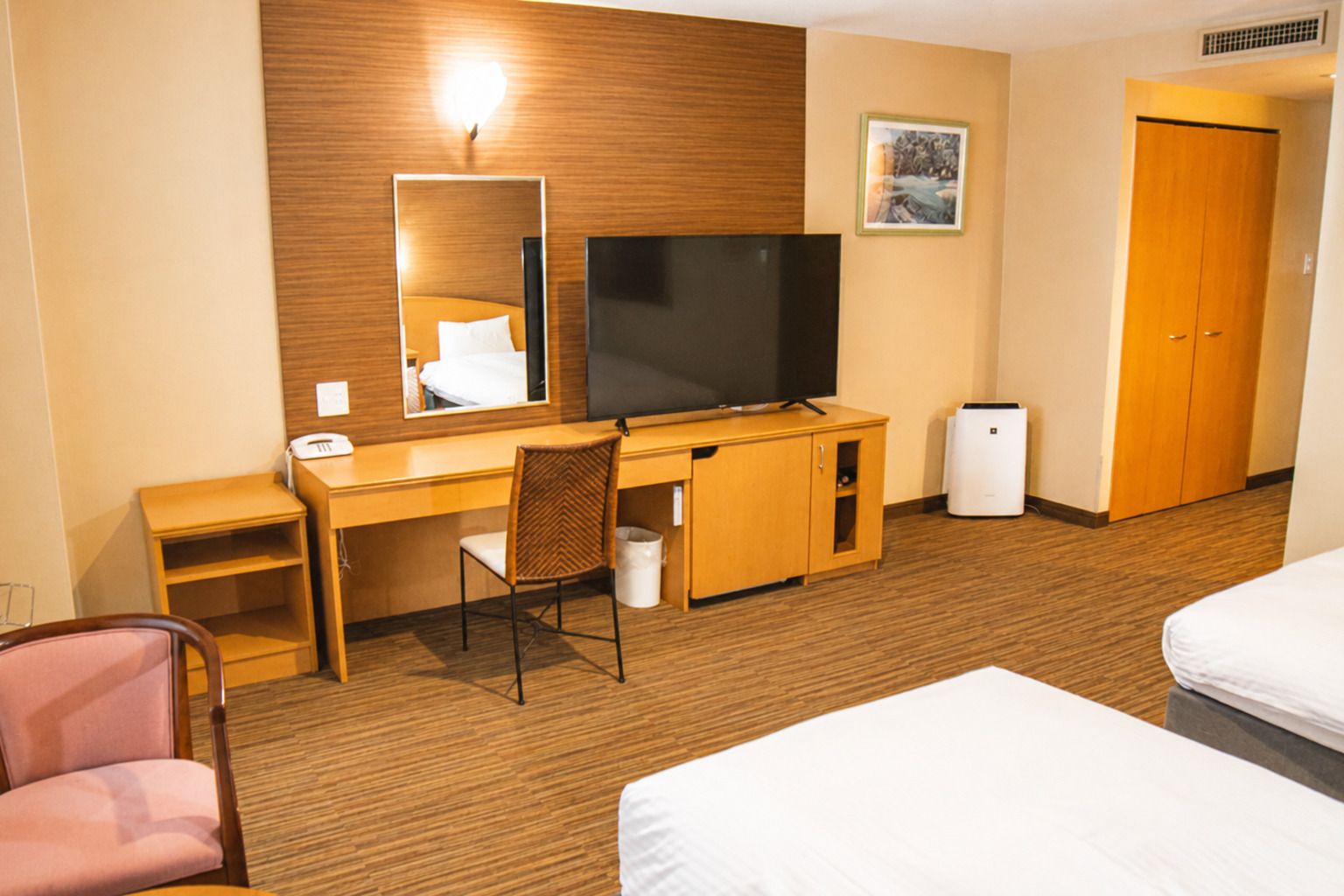 A hotel room with a wooden desk, a television, a chair, a bed, and a wardrobe.