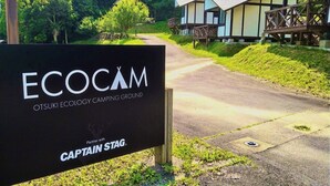 A sign for Otsuki Ecology Camping Ground with a wooden fence and a pathway leading to eco-friendly cabins.