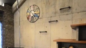 Een woonkamer met een televisie op een houten bank, een klok aan de muur en planken met planten en beeldjes.