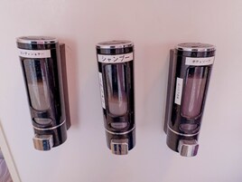 Tres recipientes cilíndricos con etiquetas, probablemente para artículos de aseo, montados en una pared.