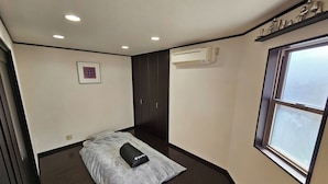A small, dimly lit room with a bed, a window, and an air conditioning unit.