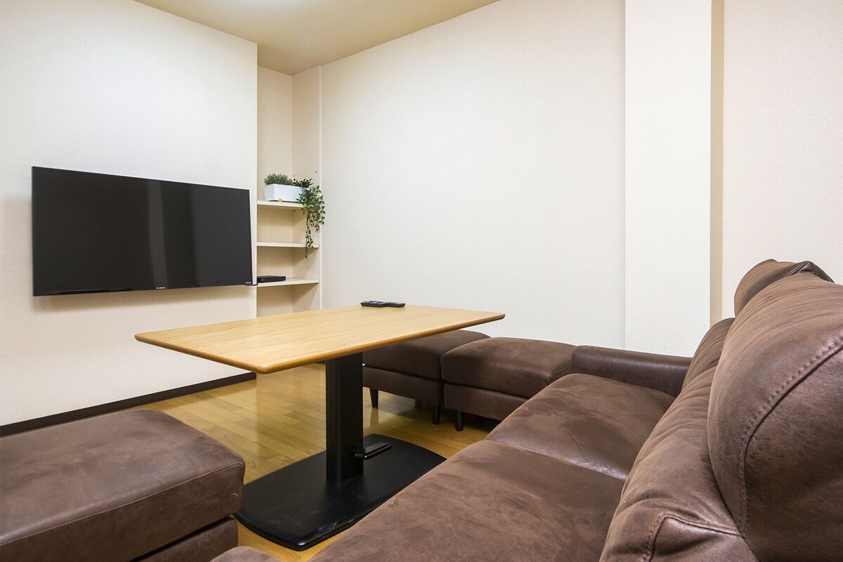 Uma sala de estar moderna com um sofá de couro marrom, uma mesa de centro de madeira, uma televisão de tela plana na parede e uma pequena planta em uma estante.