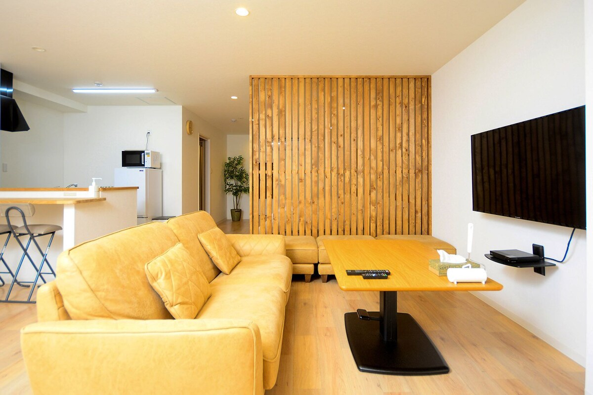 Uma sala de estar moderna com sofá, uma mesa pequena, televisão e área de cozinha ao fundo.