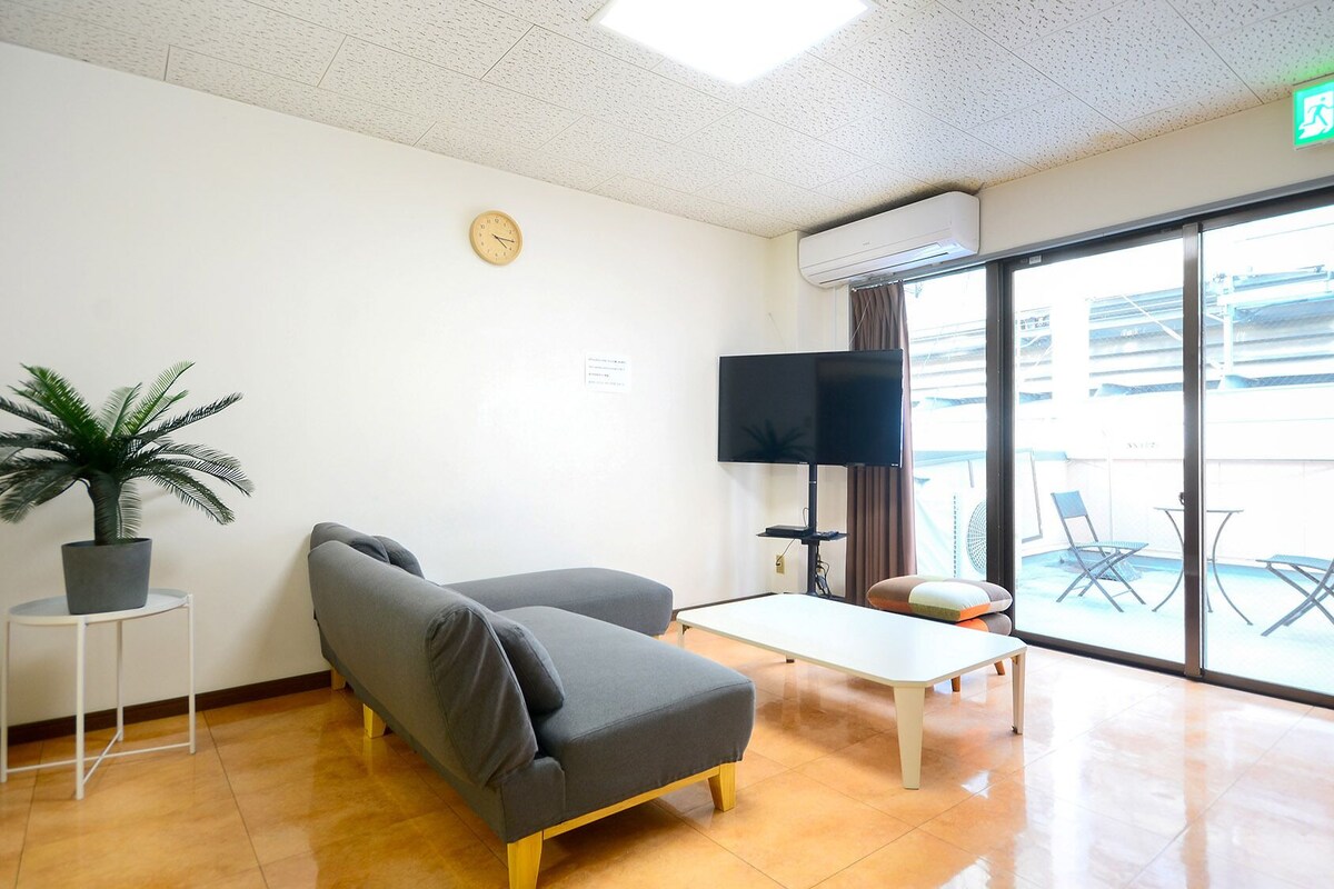 Quarto com sofá, uma mesinha, planta em vaso, televisão e porta de vidro deslizante para área externa.