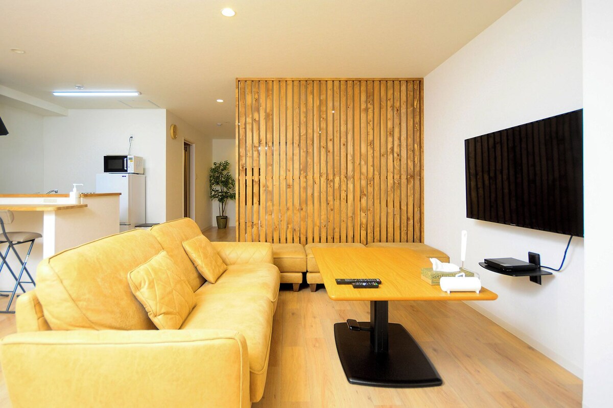 Uma sala de estar moderna com um sofá, uma divisória de madeira, uma TV e uma cozinha ao fundo.