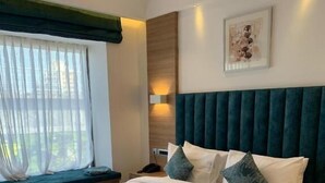 A hotel room with a large bed, a nightstand, a lamp, a window with curtains, and a wall-mounted artwork.