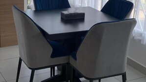 A dining area with a table and four chairs, a remote control on the table, and a view through sheer curtains.