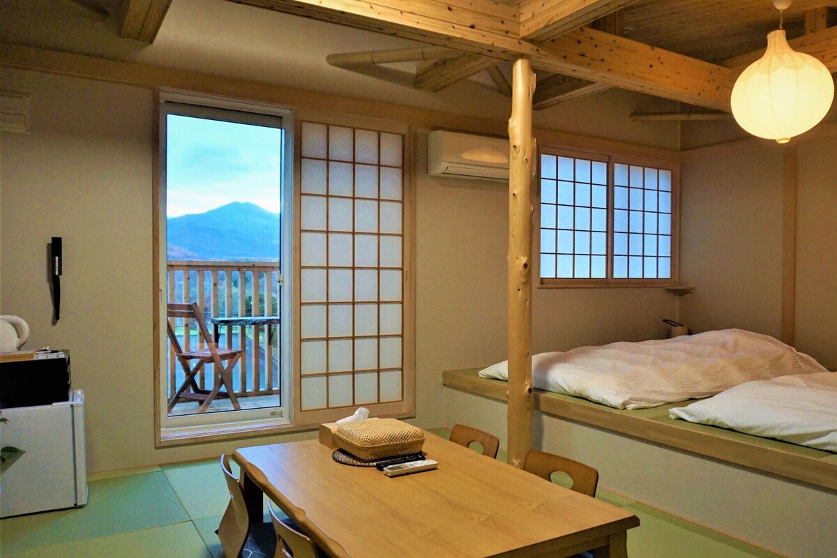 Una habitación japonesa tradicional con suelo de tatami, una mesa de madera baja, dos camas, una puerta corredera y vistas a la montaña.