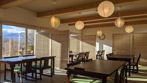 A dining area with wooden tables and chairs, large windows with blinds, and hanging paper lanterns.