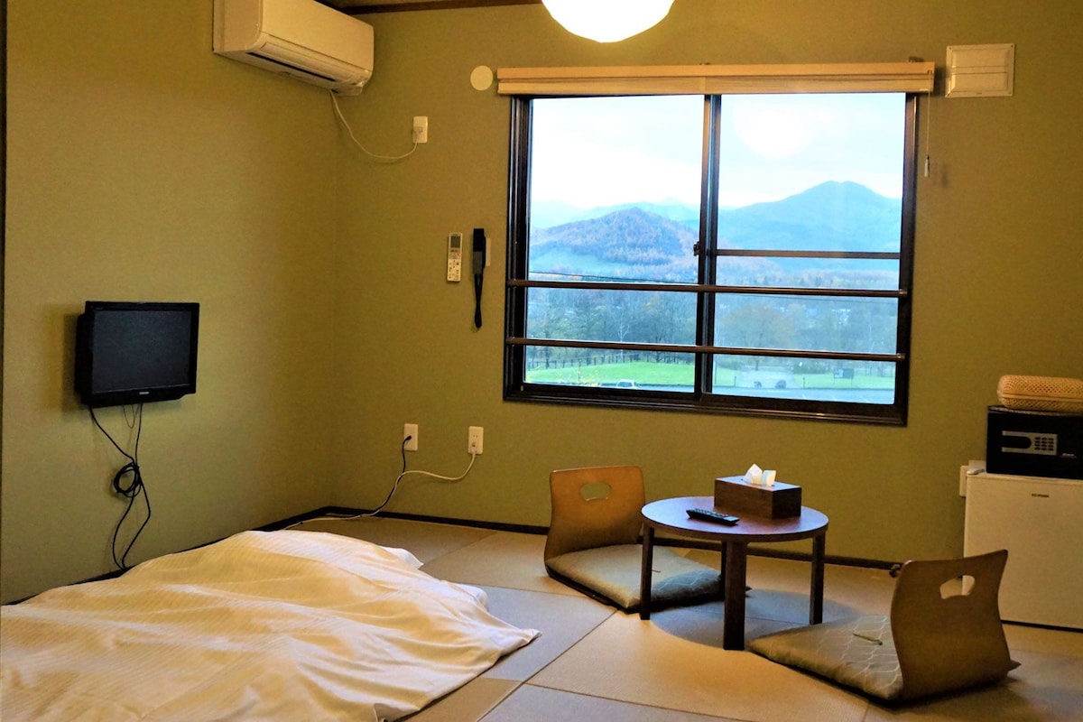 A room with tatami flooring, a bed, a small table with a remote control, a chair, a window with a view of mountains, and a small refrigerator.