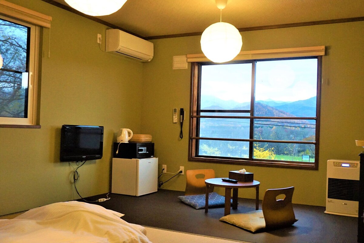 Una habitación con cama, una mesita con silla, televisión, refrigerador, microondas, cafetera, ventana con vista a la montaña y aire acondicionado.