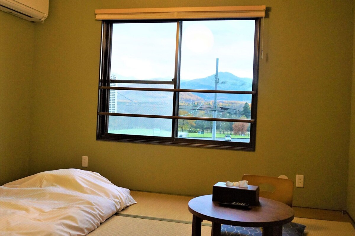 Una habitación con cama, una mesita con una caja, una ventana con vistas a montañas y un televisor montado en la pared.