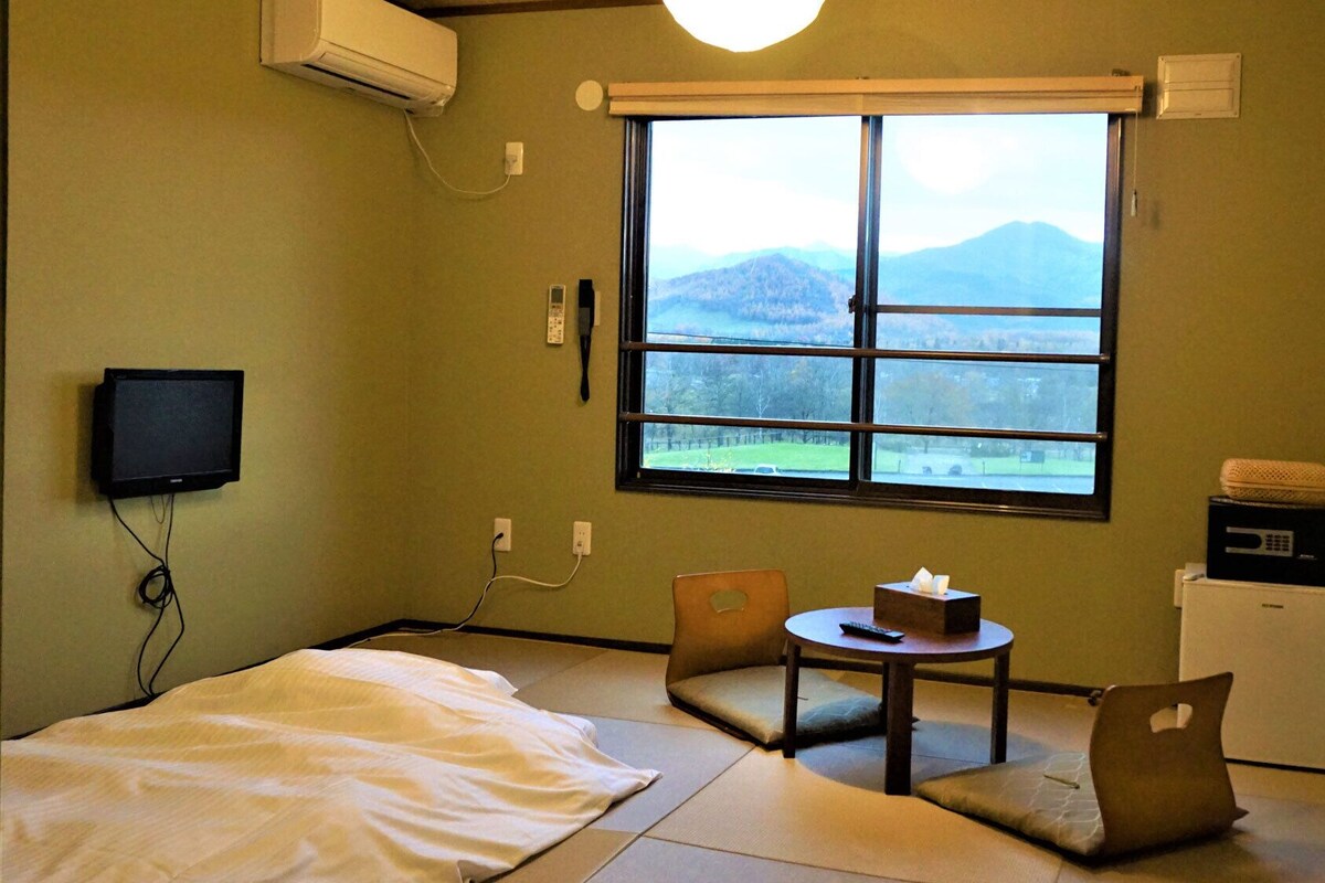 Una habitación con suelo de tatami, una cama, una mesita con un control remoto, una silla, un ventanal con vistas a montañas y un refrigerador pequeño.