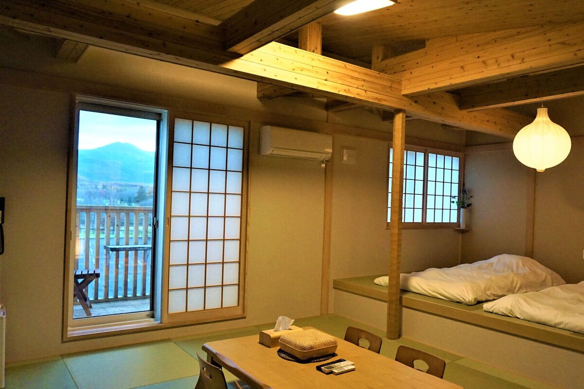 Una habitación japonesa tradicional con suelo de tatami, una mesa baja con una cesta tejida, una cama con ropa blanca y vistas a la montaña a través de un shoji.
