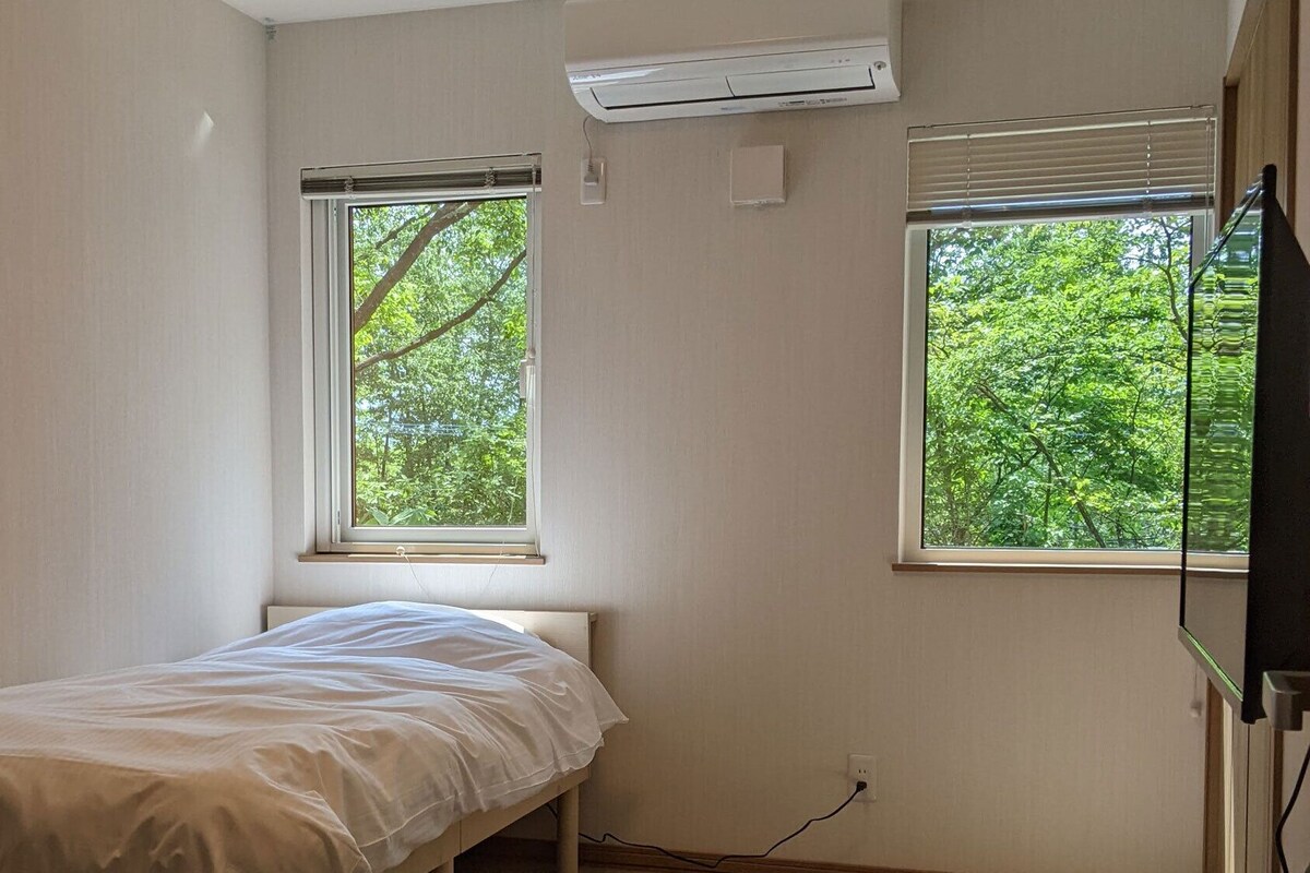 Una habitación pequeña con cama individual, dos ventanas, aire acondicionado y un enchufe.