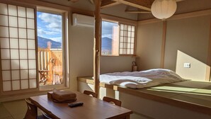 A traditional Japanese room with tatami flooring, a low bed, a wooden table with a remote control, and a view of mountains through shoji screens.